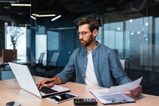 A man with glasses works at a modern office desk, focused on a laptop and documents. Nearby are a tablet, calculator, and papers, conveying a productive atmosphere.